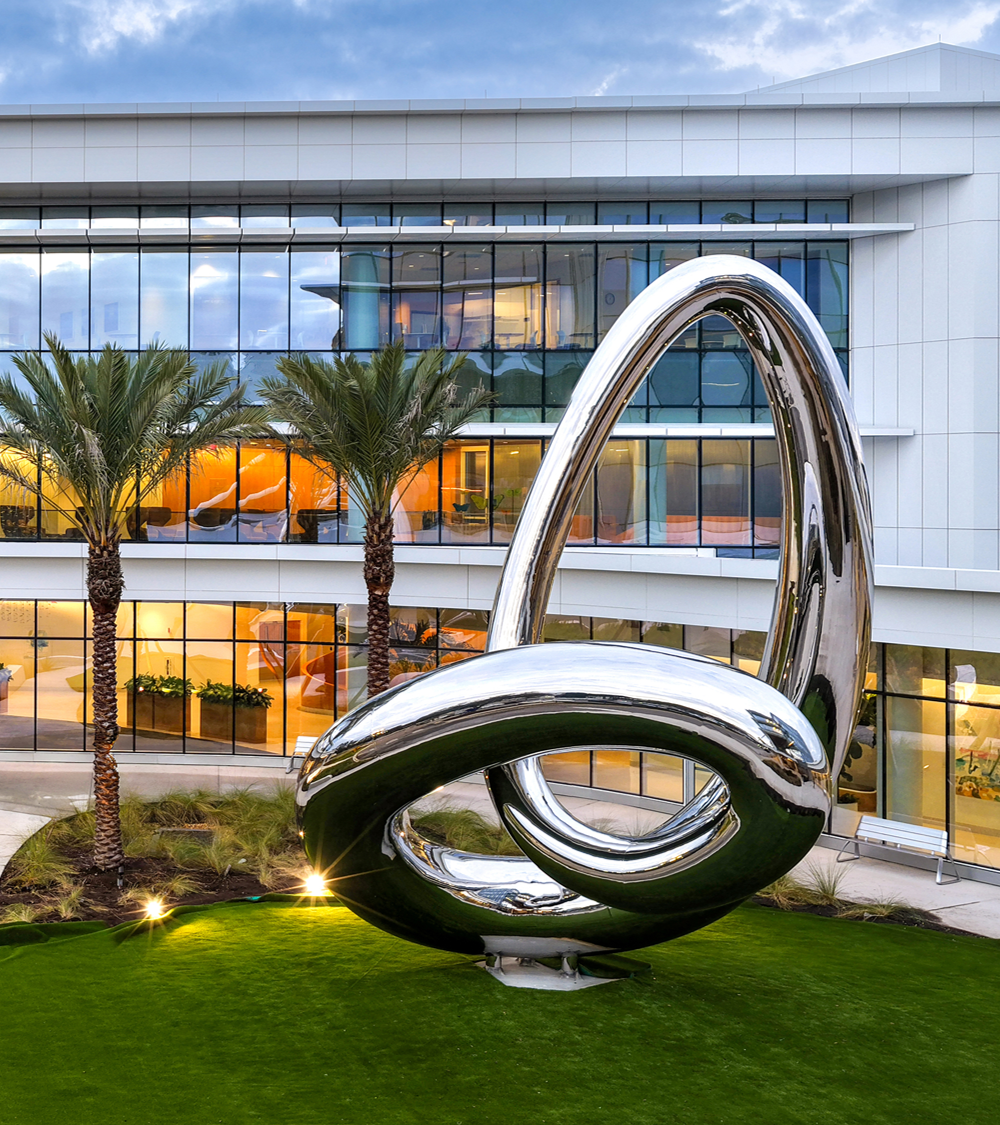 Reflective loop sculpture in a landscaped courtyard with palm trees and a glass-fronted building.